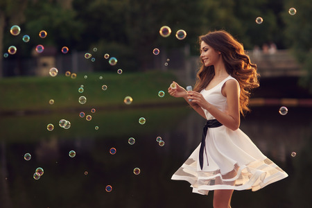 55314554 - outdoor summer portrait of young beautiful happy woman making soap bubbles in park or at nature in white dress