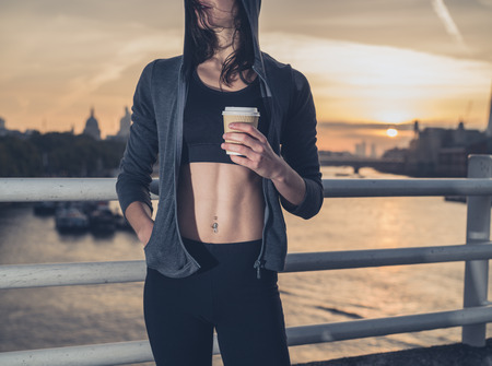 48411884 - an athletic young woman with toned abs is standing on a bridge in london at sunrise with a paper cup in her hand